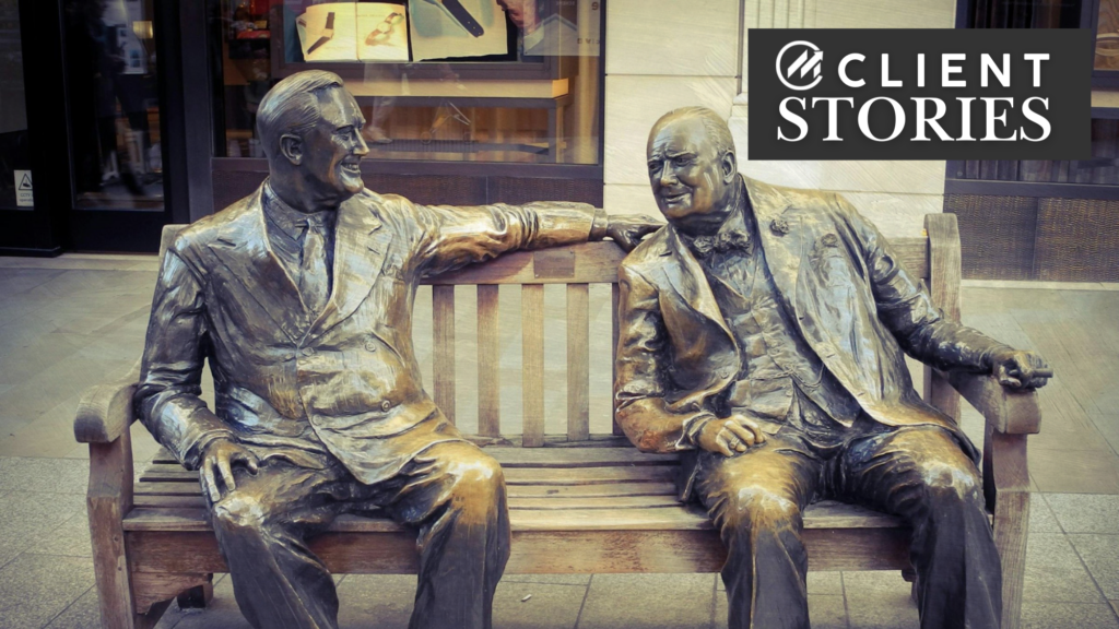 Two bronze statues of men sitting on a wooden bench on a sidewalk.