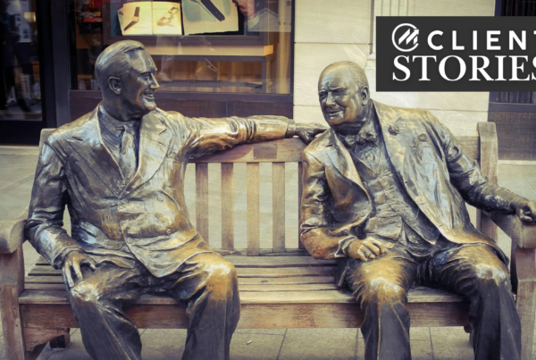 Two bronze statues of men sitting on a wooden bench on a sidewalk.